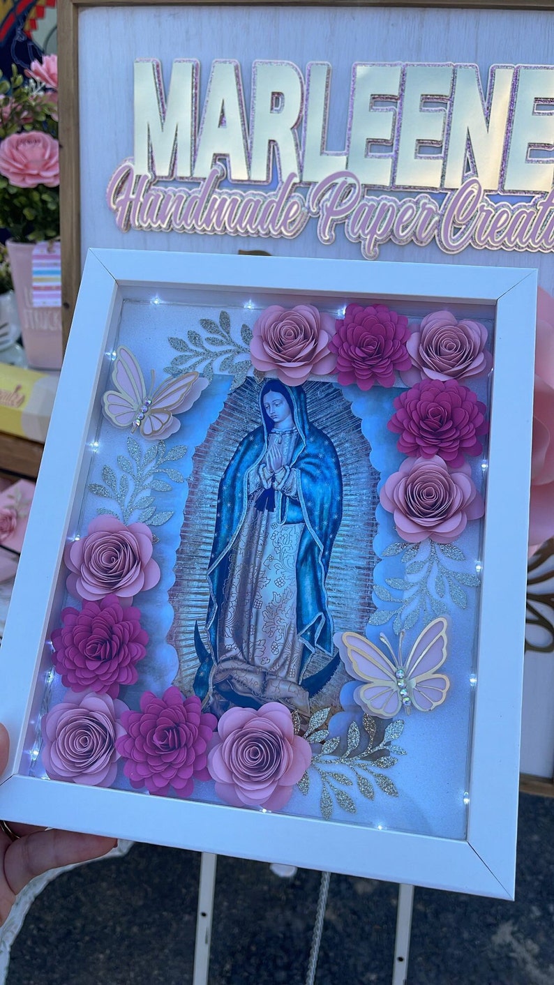 May include: A white frame with a light blue image of the Virgin Mary surrounded by pink paper flowers and butterflies. The frame has a sign above it that reads "Marleene Handmade Paper Creations".