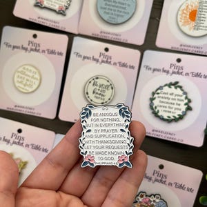 May include: A collection of enamel pins with various designs and text. One pin features the text "Be anxious for nothing, but in everything by prayer and supplication..." surrounded by floral accents. Other pins have inspirational quotes.