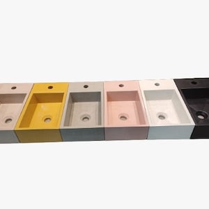 Puede incluir: Una fila de lavabos rectangulares en varios colores: beige, amarillo, gris, rosa, blanco y negro. Cada lavabo tiene una cubeta rectangular y un orificio de desagüe circular. Los lavabos son de material sólido.