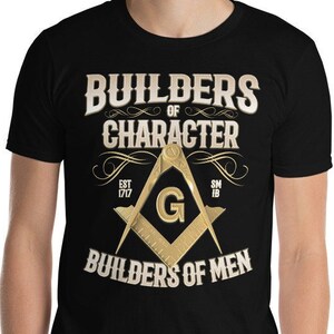 Op de afbeelding: Zwart T-shirt met een gouden vrijmetselaars symbool en de tekst "Builders of Character" en "Builders of Men".