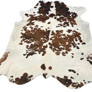 May include: White cowhide rug with brown and black spots. The rug has a natural, irregular shape.
