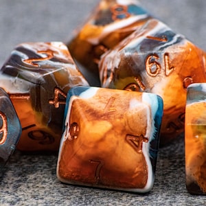May include: A set of six polyhedral dice with a marbled, multi-colored design. The dice are brown, white, and blue with copper-colored numbers. The dice are on a gray surface.