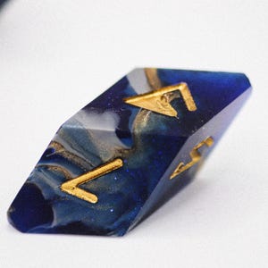 May include: A blue and white dice with gold accents. The dice is shaped like a diamond and has a swirl pattern. The number 5 is visible on the dice.