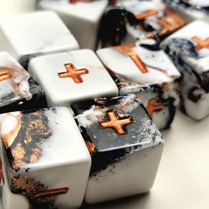 May include: Close-up of several square, decorative keycaps. The keycaps feature a white, black, and gold marbled design with a copper-colored plus sign in the center. The keycaps are likely for a computer keyboard.