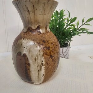 May include: A handcrafted ceramic vase with a wide, flared top and a rounded body. The vase features a speckled brown and tan glaze with white and dark brown accents. The vase is approximately 10 inches tall, suitable for displaying flowers or as a decorative piece.