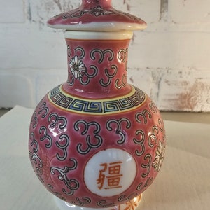 May include: A pink ceramic jar with a lid, decorated with floral and geometric patterns. The jar has a round body and a small base. The lid has a knob on top. The jar has a Chinese character that reads "福" (fú) which means "good fortune" or "happiness".