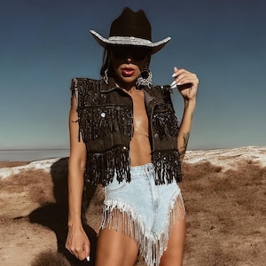 May include: A person wearing a black cowboy hat with a silver band, a black fringed vest, light blue denim shorts with silver fringe, and black cowboy boots with silver accents. The background is a desert landscape with a blue sky.