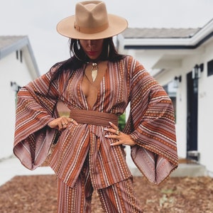 Puede incluir: Una mujer con un pantal&oacute;n de vestir a rayas marrones y blancas con un cintur&oacute;n marr&oacute;n ancho. Lleva un sombrero fedora marr&oacute;n y sandalias marrones. El atuendo tiene un estilo bohemio.