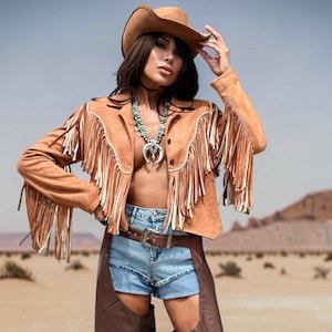 May include: A tan suede jacket with long fringe detailing, a brown cowboy hat, a turquoise squash blossom necklace, blue denim shorts with a brown leather belt, and dark brown leather chaps worn over the shorts.
