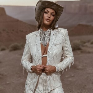 May include: A white sequined blazer and matching pants with fringe detail. The outfit is paired with a cowboy hat adorned with star-shaped studs and a large, sparkling necklace. The ensemble is set against a desert landscape.