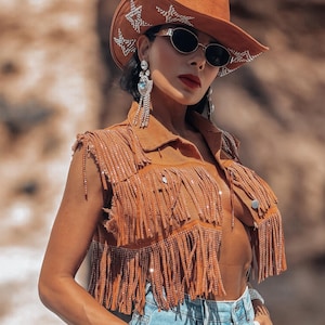 May include: A brown suede vest with fringe and rhinestone embellishments, paired with light-wash denim jeans featuring rhinestone accents. The ensemble includes a brown cowboy hat with star details and silver-toned jewelry.