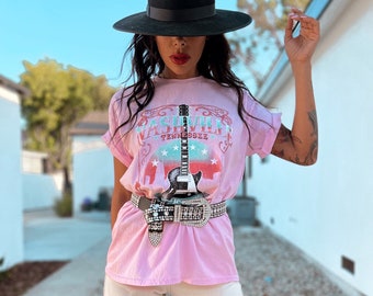 May include: A pink t-shirt with a graphic print of a guitar and the text "Nashville Tennessee" in a vintage style. The model is wearing a black hat, a rhinestone belt, and white jeans.
