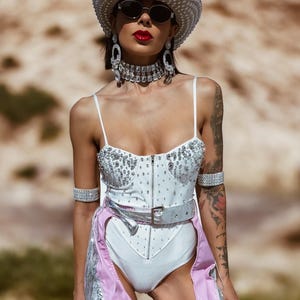 May include: A woman wearing a white bodysuit with rhinestones and pink chaps with silver fringe and stars. She is wearing a white cowboy hat, sunglasses, and silver jewellery.