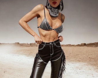 Puede incluir: Una mujer con un sujetador negro de pedrería y pantalones de cuero negro con detalles de flecos. También lleva un sombrero de vaquero plateado y un pañuelo negro.