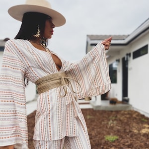 May include: A white and brown patterned pantsuit with a wide, brown woven belt. The pantsuit has a deep V-neckline and long, wide sleeves. The model is wearing a wide-brimmed, cream-colored hat.