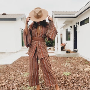 Puede incluir: Una mujer lleva un traje de pantal&oacute;n a rayas marrones y blancas con un cintur&oacute;n marr&oacute;n ancho. Lleva un sombrero marr&oacute;n y sandalias marrones. El atuendo es de estilo boho.
