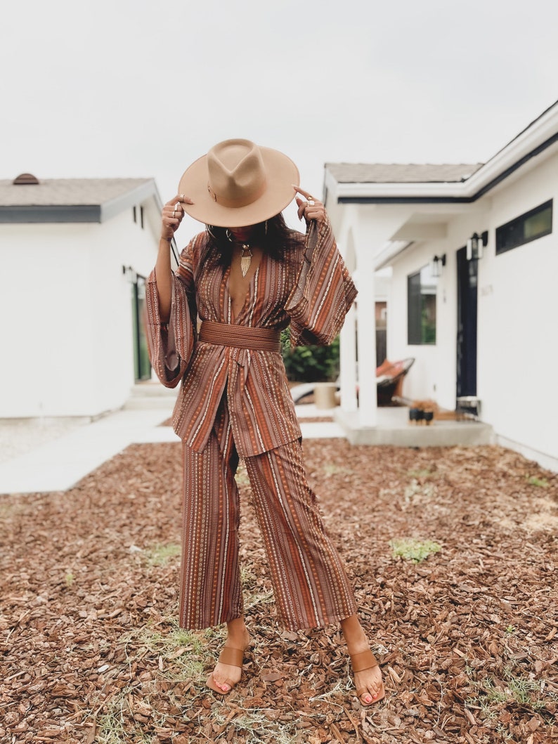 Puede incluir: Una mujer lleva un traje de pantal&oacute;n a rayas marrones y blancas con un cintur&oacute;n marr&oacute;n ancho. Lleva un sombrero marr&oacute;n y sandalias marrones. El atuendo es de estilo boho.