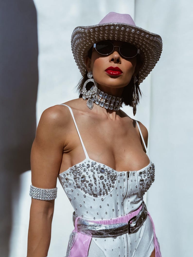May include: A woman wearing a white rhinestone-studded bodysuit with a pink and silver belt, a lavender cowboy hat with pearl embellishments, and large rhinestone earrings. She is wearing sunglasses and red lipstick.
