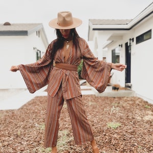 Puede incluir: Una mujer con un pantal&oacute;n de vestir a rayas marrones y blancas con un cintur&oacute;n marr&oacute;n ancho. Lleva un sombrero marr&oacute;n y sandalias marrones. El atuendo es de estilo boho.