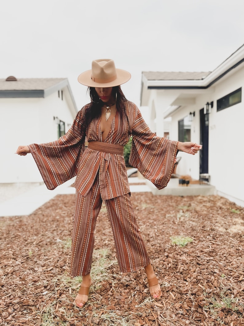 Puede incluir: Una mujer con un pantal&oacute;n de vestir a rayas marrones y blancas con un cintur&oacute;n marr&oacute;n ancho. Lleva un sombrero marr&oacute;n y sandalias marrones. El atuendo es de estilo boho.