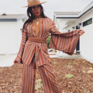 Puede incluir: Una mujer lleva un traje de pantal&oacute;n a rayas marrones y blancas con un cintur&oacute;n marr&oacute;n ancho. Lleva un sombrero marr&oacute;n y un collar de oro. El traje de pantal&oacute;n tiene un escote en V profundo y mangas largas con pantalones anchos.