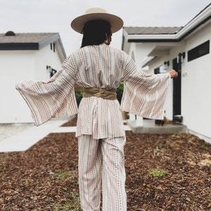 May include: A white and brown striped wide-leg pantsuit with a matching wide-brimmed hat. The pantsuit has a cinched waist with a brown woven belt.