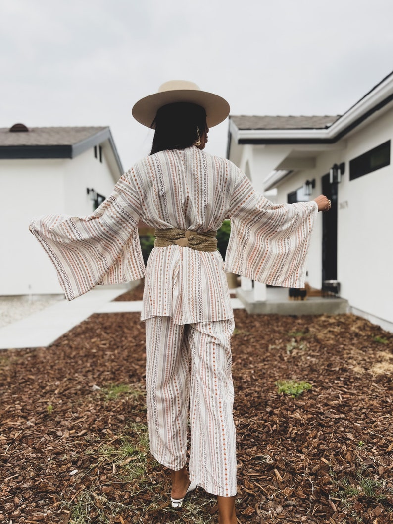 May include: A white and brown striped wide-leg pantsuit with a matching wide-brimmed hat. The pantsuit has a cinched waist with a brown woven belt.