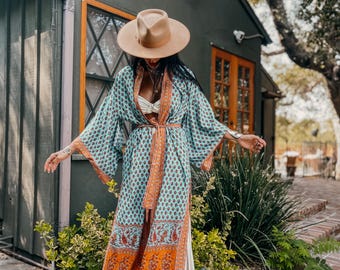 May include: A light blue patterned kimono with orange and red accents, worn with a brown hat and white pants. The kimono features a floral design and a matching belt. The outfit is completed with brown sandals.