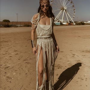 Puede incluir: Vestido de macram&eacute; color crema con aberturas altas y flecos, combinado con un pa&ntilde;uelo estampado, gafas de sol y brazaletes plateados. Una noria es visible en el fondo. El vestido tiene un cintur&oacute;n a juego.