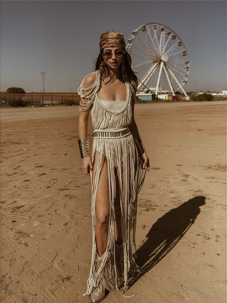 Puede incluir: Vestido de macram&eacute; color crema con aberturas altas y flecos, combinado con un pa&ntilde;uelo estampado, gafas de sol y brazaletes plateados. Una noria es visible en el fondo. El vestido tiene un cintur&oacute;n a juego.