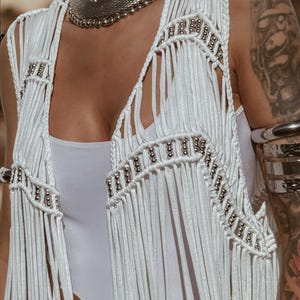May include: White fringed vest with silver bead detailing. The vest has a V-neck and silver beading along the edges. The garment is part of a bohemian style ensemble.