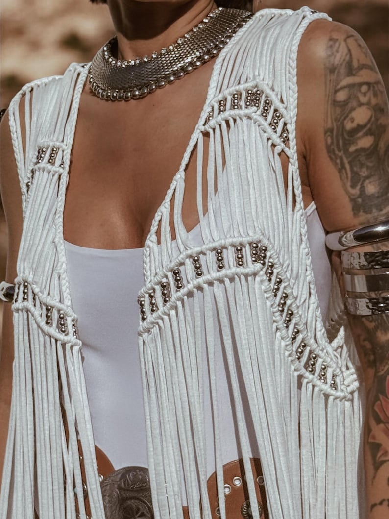 May include: White fringed vest with silver bead detailing. The vest has a V-neck and silver beading along the edges. The garment is part of a bohemian style ensemble.