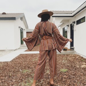Puede incluir: Una mujer con un pantal&oacute;n de vestir a rayas marrones y blancas con un cintur&oacute;n marr&oacute;n ancho. Lleva un sombrero de ala ancha y est&aacute; de pie frente a una casa.
