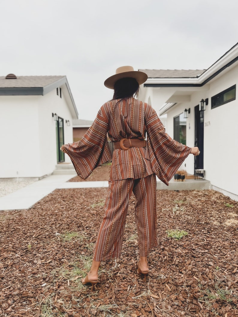 Puede incluir: Una mujer con un pantal&oacute;n de vestir a rayas marrones y blancas con un cintur&oacute;n marr&oacute;n ancho. Lleva un sombrero de ala ancha y est&aacute; de pie frente a una casa.