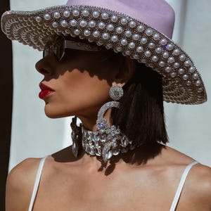 May include: A woman wearing a light purple wide-brimmed hat with a pearl trim, large silver hoop earrings, and a silver rhinestone choker.