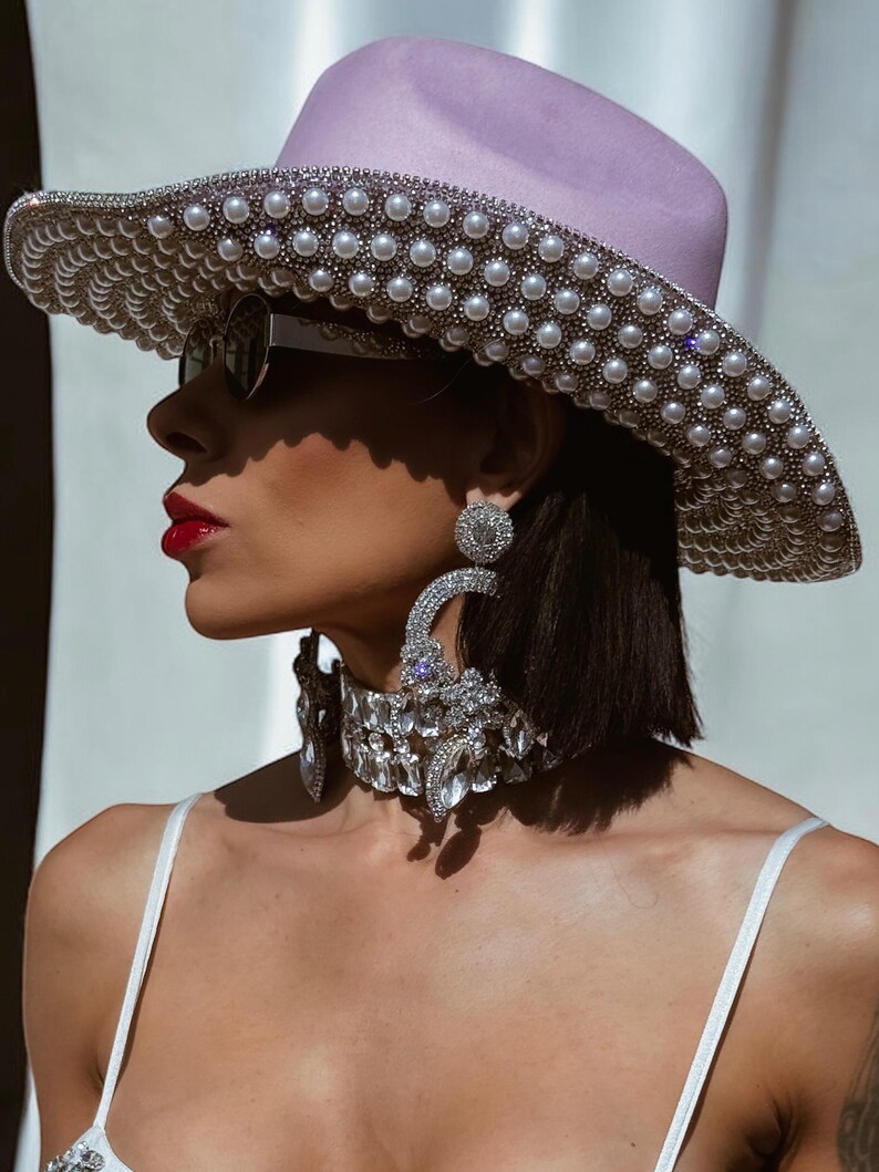 May include: A woman wearing a light purple wide-brimmed hat with a pearl trim, large silver hoop earrings, and a silver rhinestone choker.