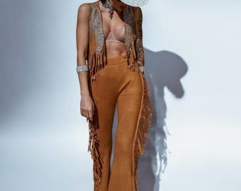 May include: A woman wearing a brown suede fringe pantsuit with a white cowboy hat. The pantsuit has a vest with rhinestones and fringe.