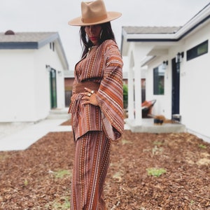 Puede incluir: Una mujer con un traje de pantal&oacute;n a rayas marrones y blancas con un sombrero de ala ancha. El traje de pantal&oacute;n tiene un patr&oacute;n geom&eacute;trico y el sombrero es de color marr&oacute;n claro. La mujer est&aacute; de pie sobre un lecho de mantillo marr&oacute;n.
