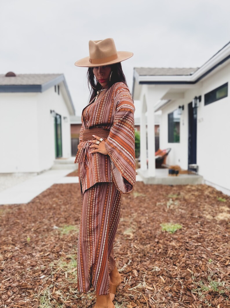 Puede incluir: Una mujer con un traje de pantal&oacute;n a rayas marrones y blancas con un sombrero de ala ancha. El traje de pantal&oacute;n tiene un patr&oacute;n geom&eacute;trico y el sombrero es de color marr&oacute;n claro. La mujer est&aacute; de pie sobre un lecho de mantillo marr&oacute;n.