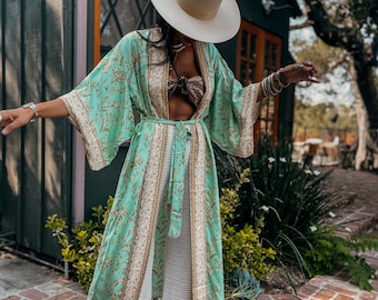 Puede incluir: Un kimono verde claro con estampado floral, mangas anchas y cinturón a juego. Se combina con pantalones palazzo blancos a rayas y un sombrero crema de ala ancha. El conjunto se complementa con múltiples pulseras.
