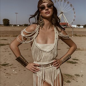 Puede incluir: Una mujer con un body de macram&eacute; color crema con flecos, un cintur&oacute;n a juego y brazaletes. Tambi&eacute;n lleva un pa&ntilde;uelo en la cabeza estampado, gafas de sol y pendientes dorados grandes. Una noria es visible en el fondo.
