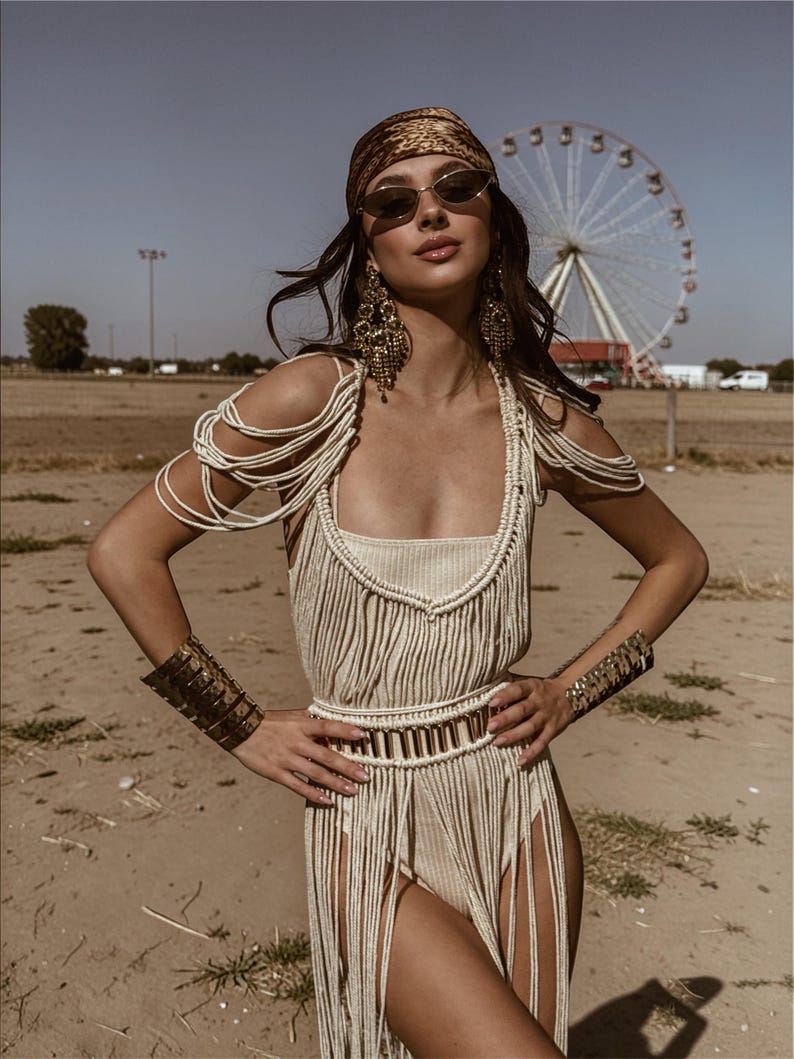 Puede incluir: Una mujer con un body de macram&eacute; color crema con flecos, un cintur&oacute;n a juego y brazaletes. Tambi&eacute;n lleva un pa&ntilde;uelo en la cabeza estampado, gafas de sol y pendientes dorados grandes. Una noria es visible en el fondo.