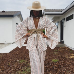 May include: A white and brown striped wide-leg pantsuit with a wide, flowing kimono-style top. The outfit is accessorized with a wide straw belt and a cream-colored fedora hat.