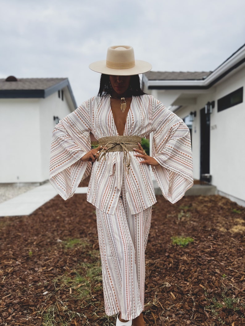 May include: A white and brown striped wide-leg pantsuit with a wide, flowing kimono-style top. The outfit is accessorized with a wide straw belt and a cream-colored fedora hat.