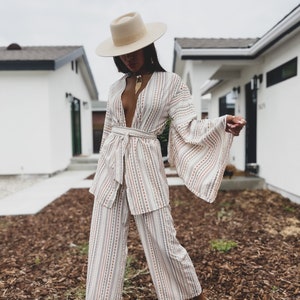 May include: A woman wearing a white and brown striped pantsuit with a wide-brimmed hat and white sandals. The pantsuit has a deep V-neckline and a belt at the waist. The woman is standing in front of a house with a white exterior.