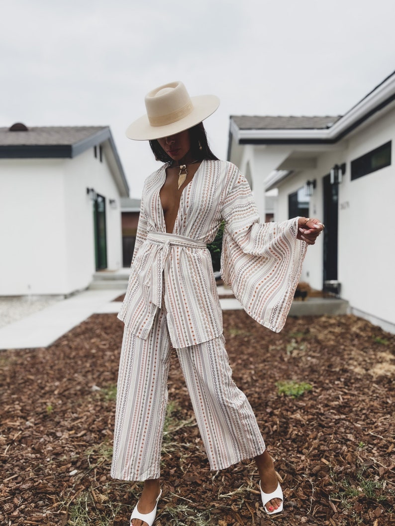 May include: A woman wearing a white and brown striped pantsuit with a wide-brimmed hat and white sandals. The pantsuit has a deep V-neckline and a belt at the waist. The woman is standing in front of a house with a white exterior.