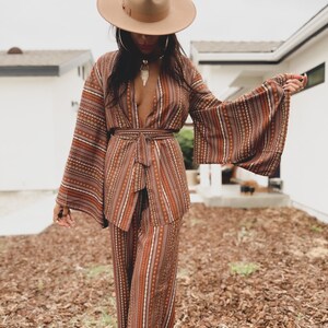 Puede incluir: Una mujer lleva un traje de pantal&oacute;n marr&oacute;n y blanco con estampado y un sombrero marr&oacute;n de ala ancha. El traje de pantal&oacute;n tiene un escote en V profundo y mangas largas y fluidas. La mujer est&aacute; de pie en un patio con mantillo marr&oacute;n.