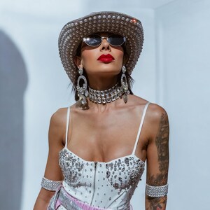 May include: A woman wearing a white bodysuit with rhinestones, a silver belt, and a silver cowboy hat with rhinestones. She is also wearing silver earrings, a silver choker, and silver bracelets. She has red lipstick and is looking up.