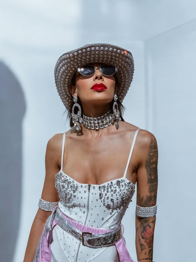 May include: A woman wearing a white bodysuit with rhinestones, a silver belt, and a silver cowboy hat with rhinestones. She is also wearing silver earrings, a silver choker, and silver bracelets. She has red lipstick and is looking up.