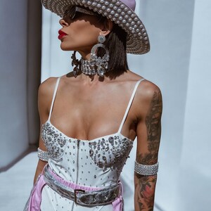 May include: A white bodysuit with a corset style bodice and a silver belt. The bodysuit is covered in rhinestones. The model is wearing a pink cowboy hat with pearl embellishments and large rhinestone earrings.